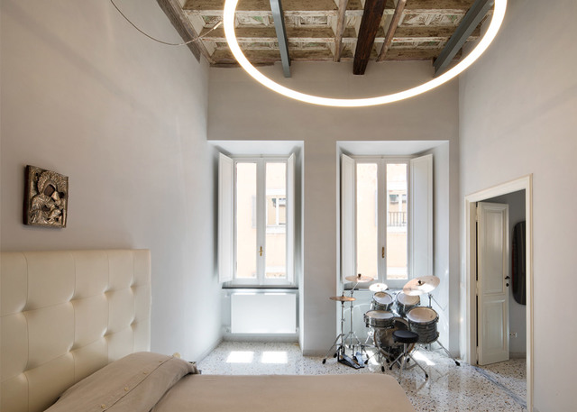 Lampada a sospensione circolare LED Artemide che illumina una camera luminosa con travi in legno a vista, pareti bianche e una batteria vicino a finestre alte.