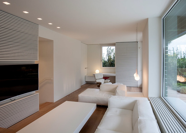 Modern white living room with recessed ceiling lights, two slim pendant lamps, white sofa and ottoman, large window with garden view