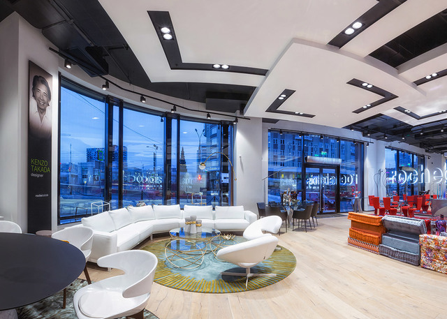 Lobby di hotel moderno con faretti a soffitto incassati, divano bianco curvo, sedie di design e finestre dal pavimento al soffitto con vista su una strada cittadina al crepuscolo.