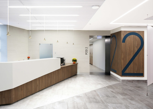 Area reception sanitaria moderna con scrivania curva bianca, dettagli in legno, luci lineari a soffitto e grande numero 2 che indica il Pod 2.