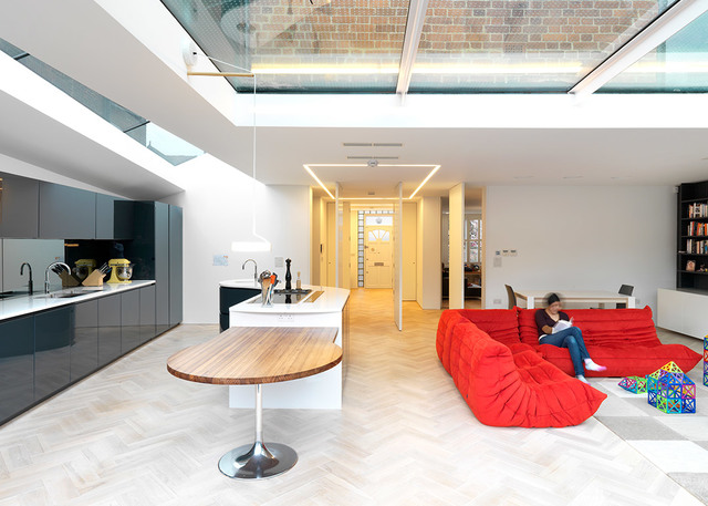 Spazio living open space moderno con lucernario, luci a soffitto LED lineari incassate, isola cucina, pavimento a spina di pesce e zona divano rosso.