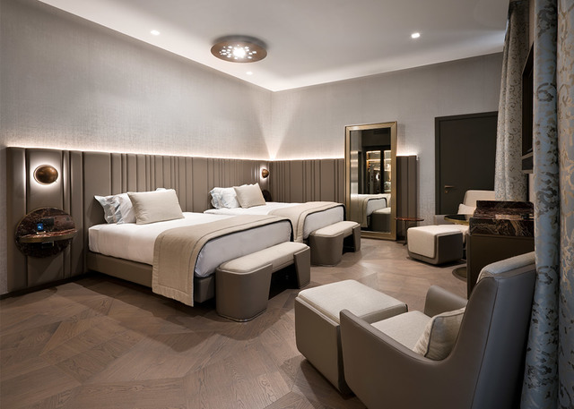 Elegant hotel room with two beds, herringbone wood floor, recessed ceiling lights, circular wall sconces, and a flush ceiling fixture.
