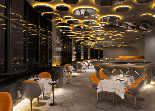 Interno di ristorante elegante con soffitto scenografico di elementi circolari Artemide ambrati, sedie arancioni, tavoli con tovaglia bianca, vista città notturna