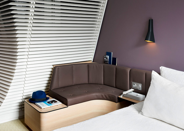 Camera d'albergo con lampada da parete conica nera Artemide sopra un divano curvo in pelle marrone accanto a un letto bianco e veneziane