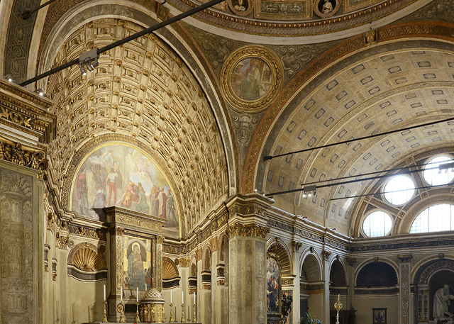 Interior of a Renaissance church with ornate gilded vaulted ceilings, frescoed arches, and an altar, lit by track lighting on ceiling rails.