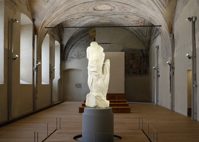 Michelangelo's unfinished Rondanini Pietà sculpture displayed on a pedestal in a vaulted museum hall with Artemide spotlights