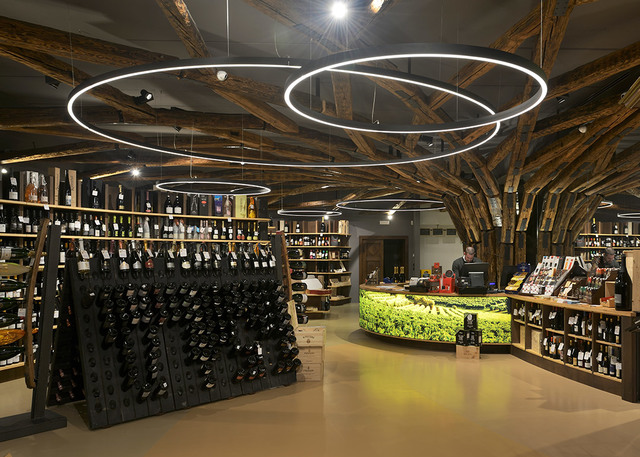 Interno di enoteca con lampade a sospensione circolari LED Artemide Halo appese a soffitto con travi rustiche sopra espositori di bottiglie.
