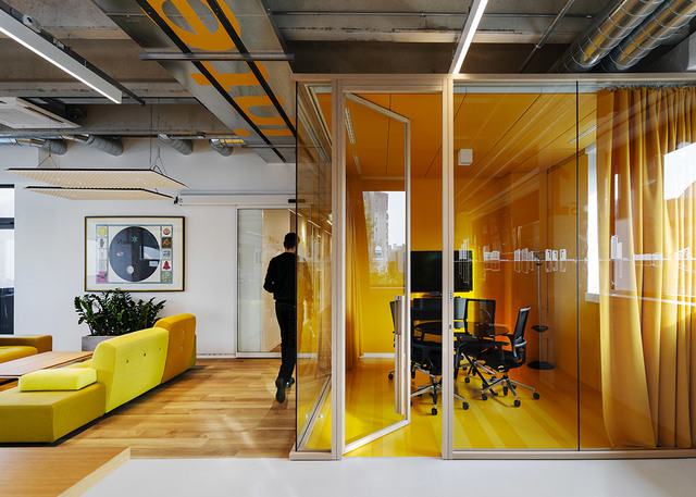 Ufficio moderno con sala riunioni a pareti di vetro giallo, luci lineari a soffitto e area lounge aperta con divano modulare giallo.