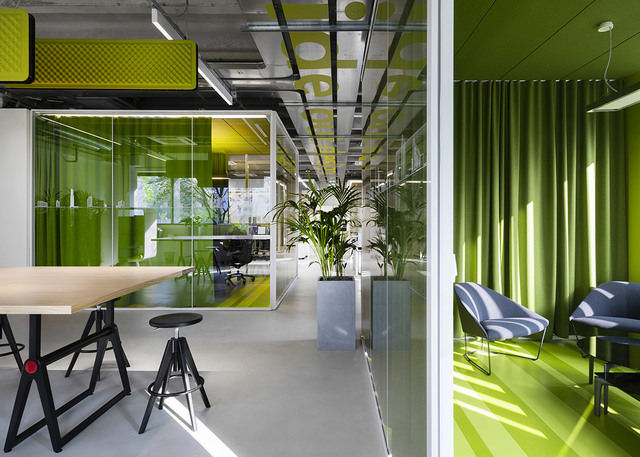 Ufficio open space moderno con vivaci accenti verdi, pareti in vetro, illuminazione a sospensione su soffitto a vista, postazioni di lavoro e rigogliose piante