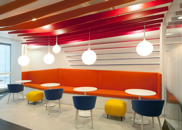 Lounge moderno con cinque lampade a sospensione sferiche bianche sopra un divano arancione, sedie colorate e pouf gialli sotto un soffitto a listelli rossi