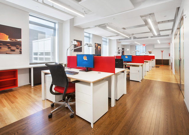 Ufficio open space moderno con postazioni bianche, divisori rossi, lampade da tavolo e luci lineari a soffitto che illuminano uno spazio con pavimento in legno.