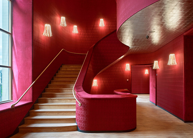 Elegant red interior with curved staircase featuring wall-mounted fringe-shade sconces casting warm light against crimson walls.