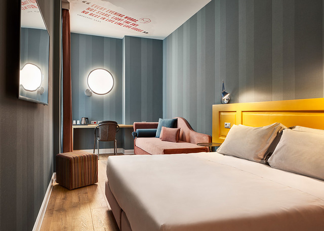 Hotel room with two round white wall sconces above a desk, yellow headboard bed, pink armchair, and striped blue-grey walls