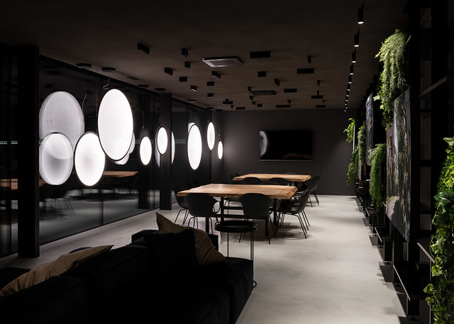 Lounge moderno scuro con grandi lampade a sospensione circolari luminose Artemide, parete verde viva, tavoli in legno e faretti a binario a soffitto