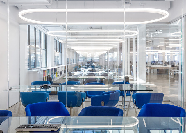 Modern office with glass-top tables, blue chairs, and Artemide oval ring pendant lights aligned along the ceiling.
