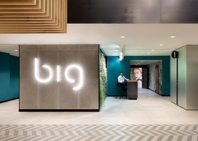 Modern office lobby with illuminated 'big' logo sign, living green wall, receptionist at desk, teal walls, and recessed ceiling lights
