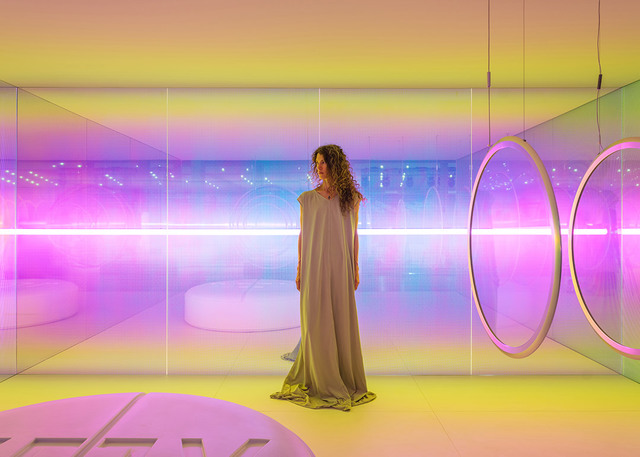 Donna in una stanza a specchi con illuminazione a gradiente rosa e giallo vibrante, lampade a sospensione circolari e strisce di luce al neon.