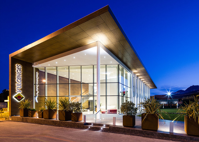 Esterno di un edificio commerciale moderno al crepuscolo, con grande facciata in vetro, tetto a sbalzo con illuminazione incassata calda e ingresso paesaggistico.
