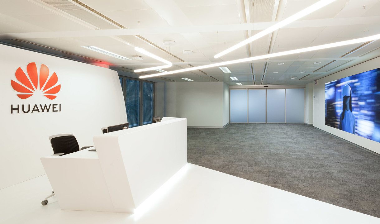 Reception ufficio Huawei con scrivania bianca, lampade lineari LED Artemide a soffitto che illuminano un moderno spazio aperto con pavimento in moquette.
