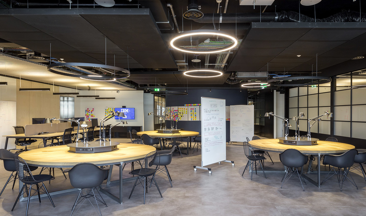 Modern collaborative workspace with round wooden tables, black chairs, and Artemide circular LED pendant lights illuminating the open office.