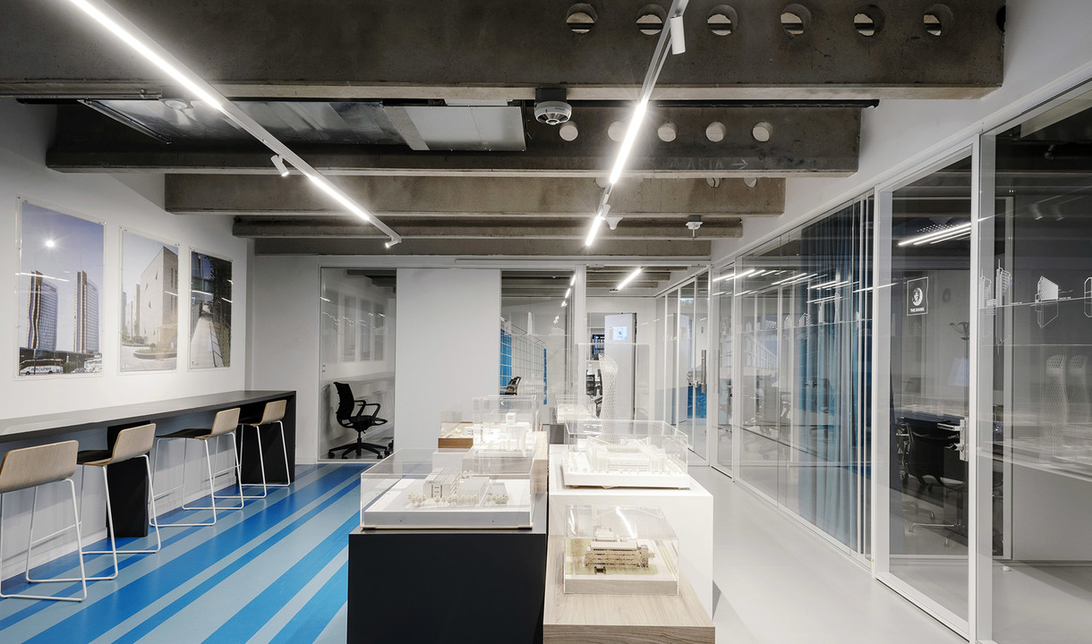 Ufficio moderno con lampade lineari LED Artemide a soffitto che illuminano modelli architettonici su tavoli espositivi e spazi di lavoro con pareti in vetro.