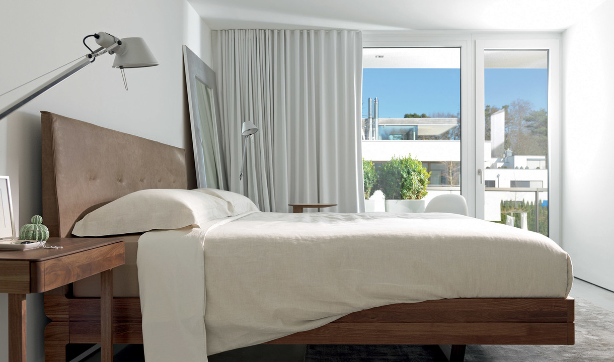 Camera da letto moderna con lampada da terra Artemide Tolomeo in argento accanto a un letto in legno con biancheria neutra, grandi finestre con tende bianche