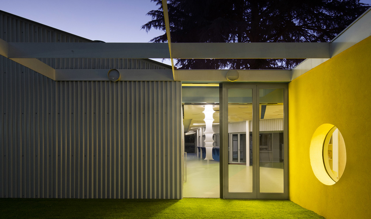 Esterno di edificio moderno al crepuscolo con parete gialla, finestra circolare e lampada colonnare bianca luminosa visibile attraverso porte in vetro.