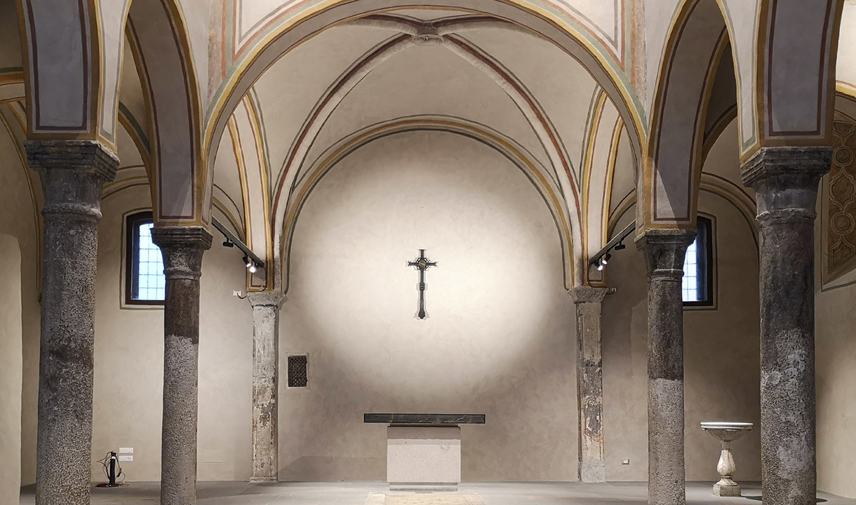 Interno di cappella storica con soffitti a volta, colonne in pietra e crocifisso sull'abside, illuminato da faretti discreti