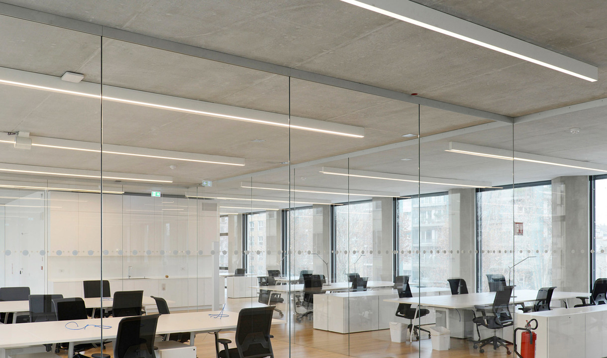 Ufficio open space moderno con lampade a sospensione LED lineari appese a soffitto in cemento a vista, che illuminano postazioni di lavoro divise da pareti in vetro.