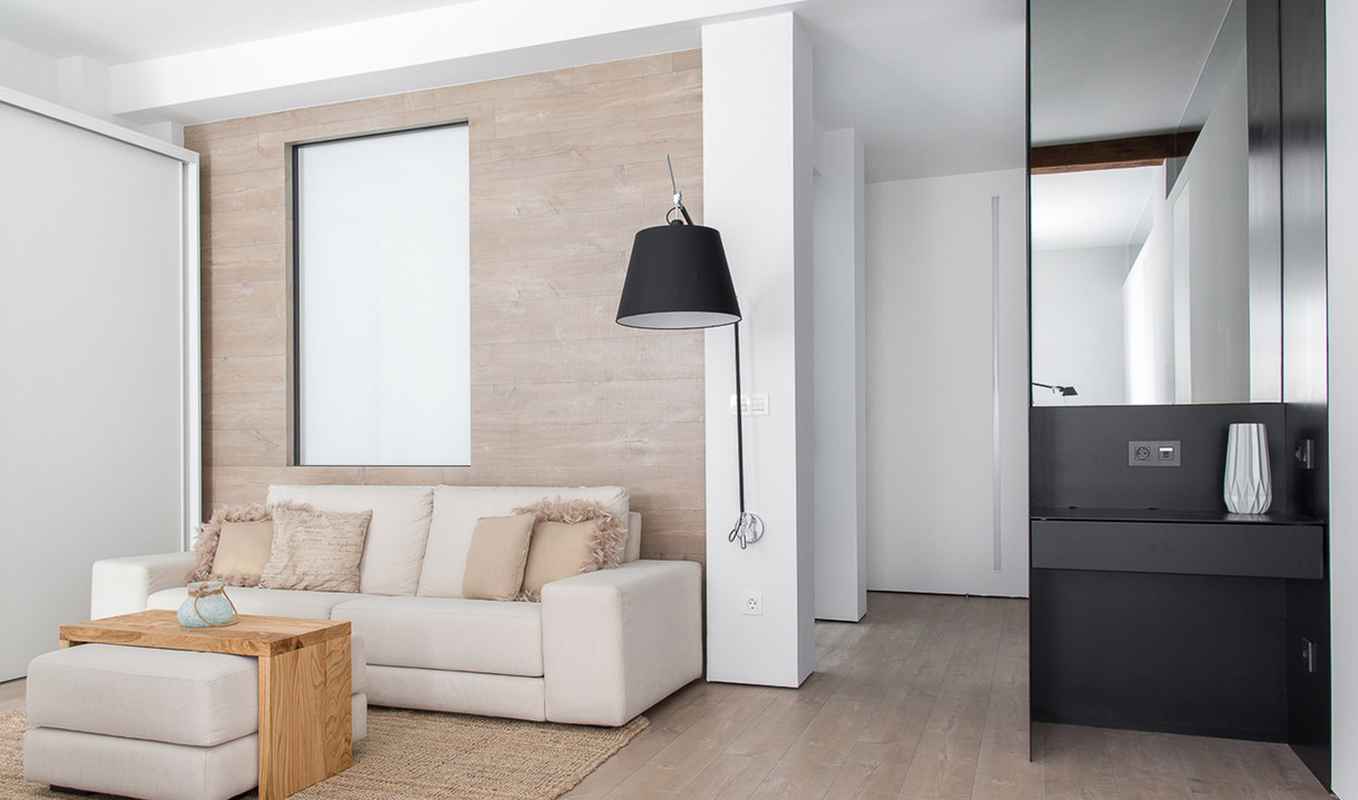Soggiorno moderno con lampada da terra conica nera accanto a un divano bianco, toni neutri, dettagli in legno e uno scorcio del bagno.
