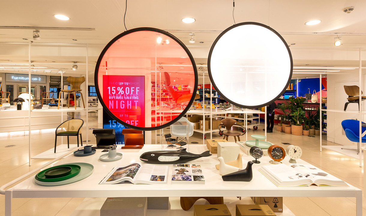 Interno di un negozio di design con due grandi lampade a sospensione circolari, una rossa e una bianca, sopra un tavolo espositivo con oggetti di design.