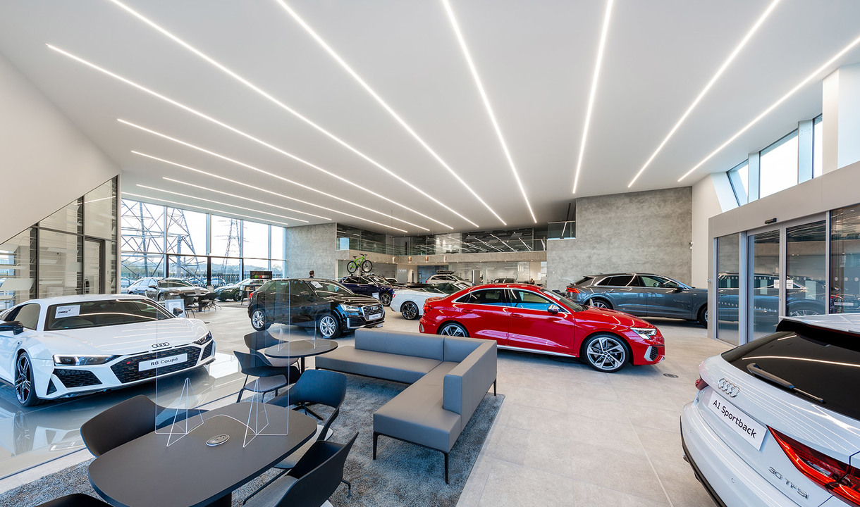 Showroom Audi con luci lineari a LED a soffitto che illuminano i veicoli esposti su pavimento lucido con area lounge
