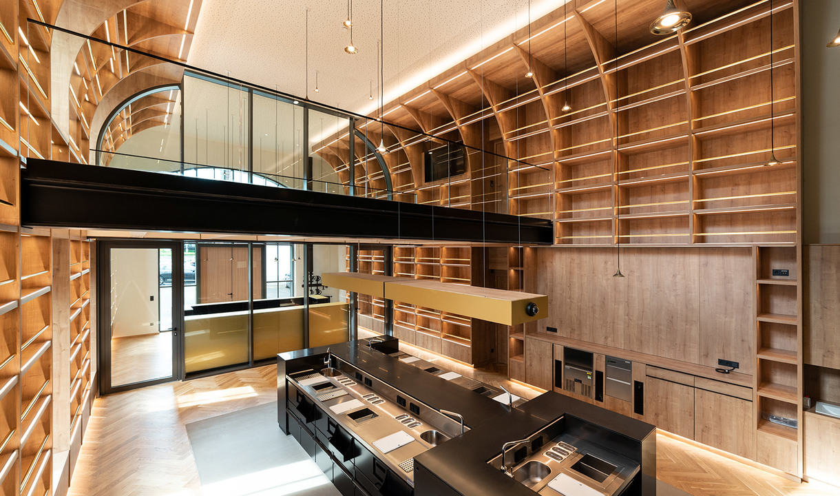 Interno di biblioteca a doppia altezza con scaffalature in legno caldo, pavimento a spina di pesce, luci a sospensione e isola cucina scura sotto un soppalco.