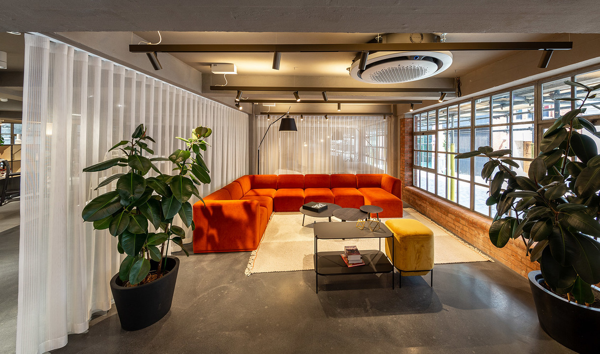 Modern lounge with orange sectional sofa, yellow ottoman, arc floor lamp, sheer curtains, concrete floor, and large industrial windows.