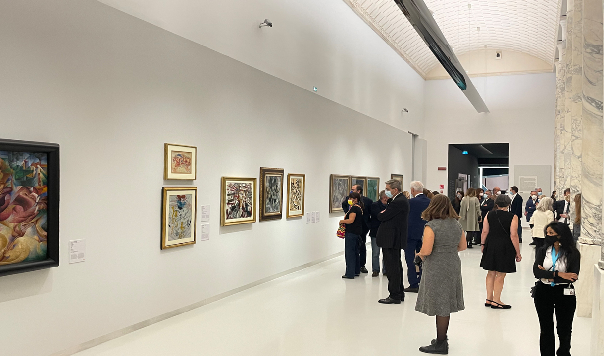 Galleria museale con visitatori che osservano dipinti colorati su pareti bianche, illuminati da faretti a soffitto incassati.