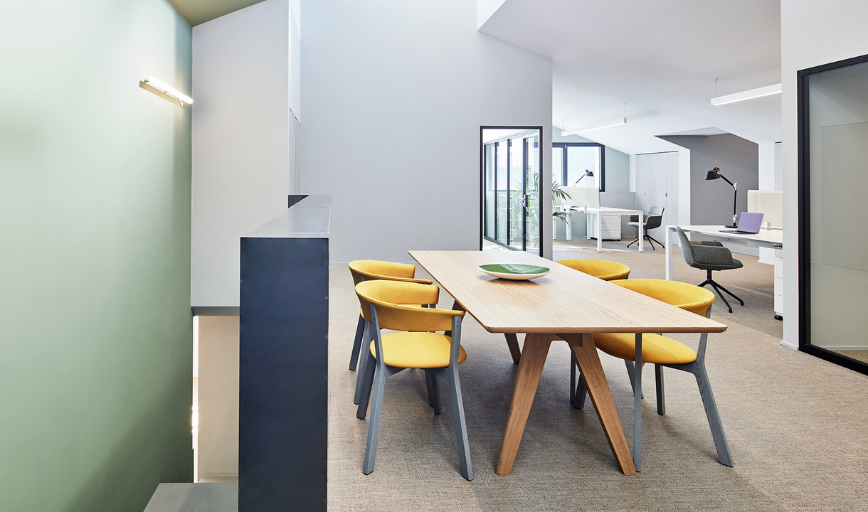 Modern office meeting area with wooden table, yellow chairs, and a slim wall-mounted linear light fixture on a sage green wall.
