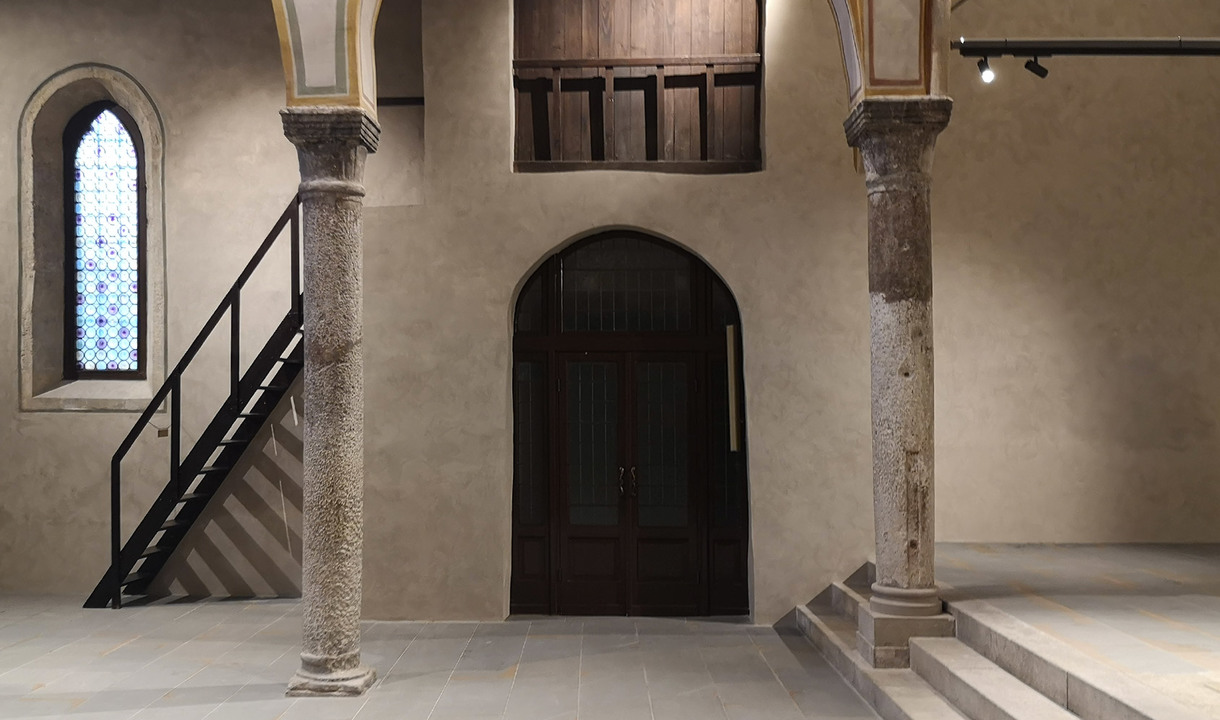 Interno storico con colonne in pietra, porta ad arco in legno, vetrata colorata, scala nera e faretti su binario Artemide a soffitto.