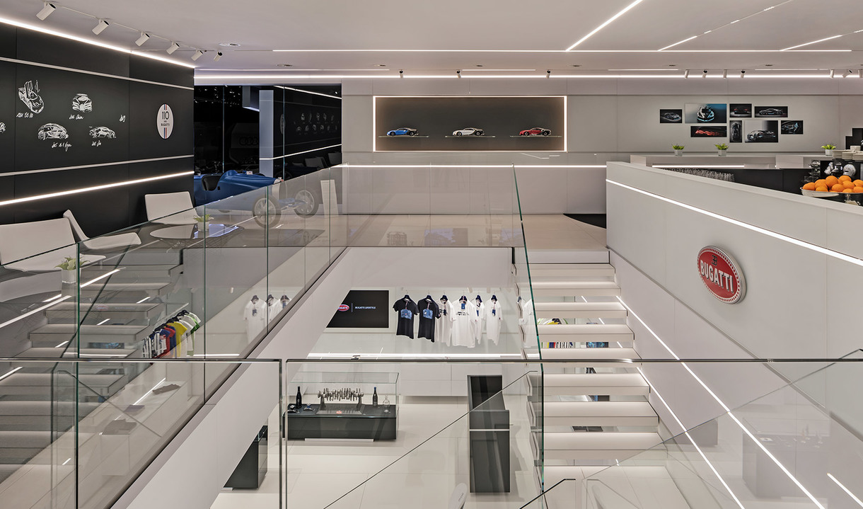 Interno showroom Bugatti su due livelli, design minimalista bianco, illuminazione LED lineare, ringhiere in vetro ed espositori di merchandising di lusso