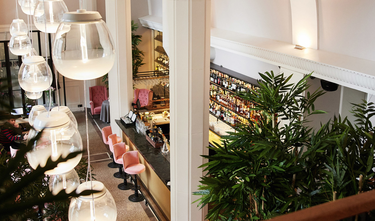Elegant restaurant interior with large glass globe pendant lights hanging over a bar area with pink stools and lush green plants.