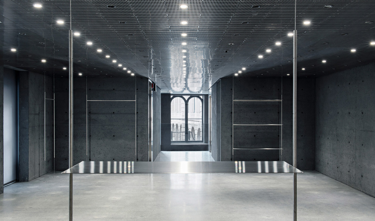 Minimalist concrete interior with recessed ceiling spotlights illuminating a symmetrical space with glass partitions and arched window.