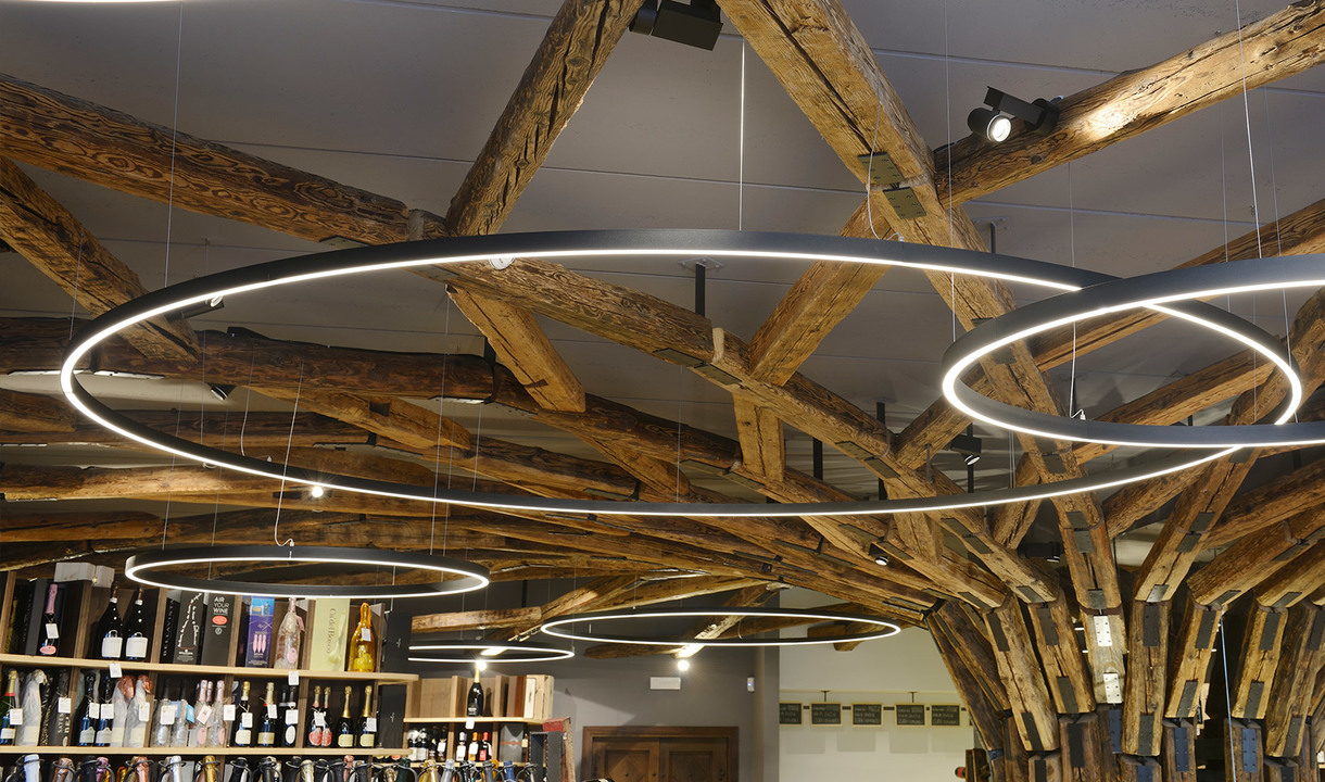 Più anelli a sospensione LED circolari Artemide di diverse dimensioni sospesi sopra un'enoteca con soffitto a travi in legno rustico.