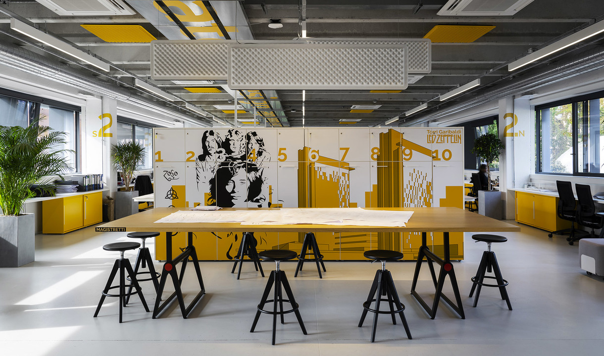 Spazio di lavoro moderno con accenti gialli, grande tavolo da lavoro, sgabelli neri, lampade a sospensione rettangolari e parete con murales grafico.