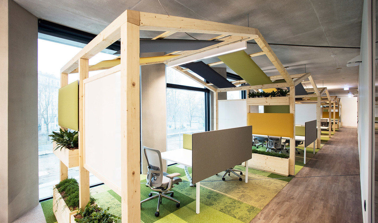 Modern open office with wooden-framed workstations, green acoustic panels on ceiling, colorful rugs, and ergonomic chairs along a corridor