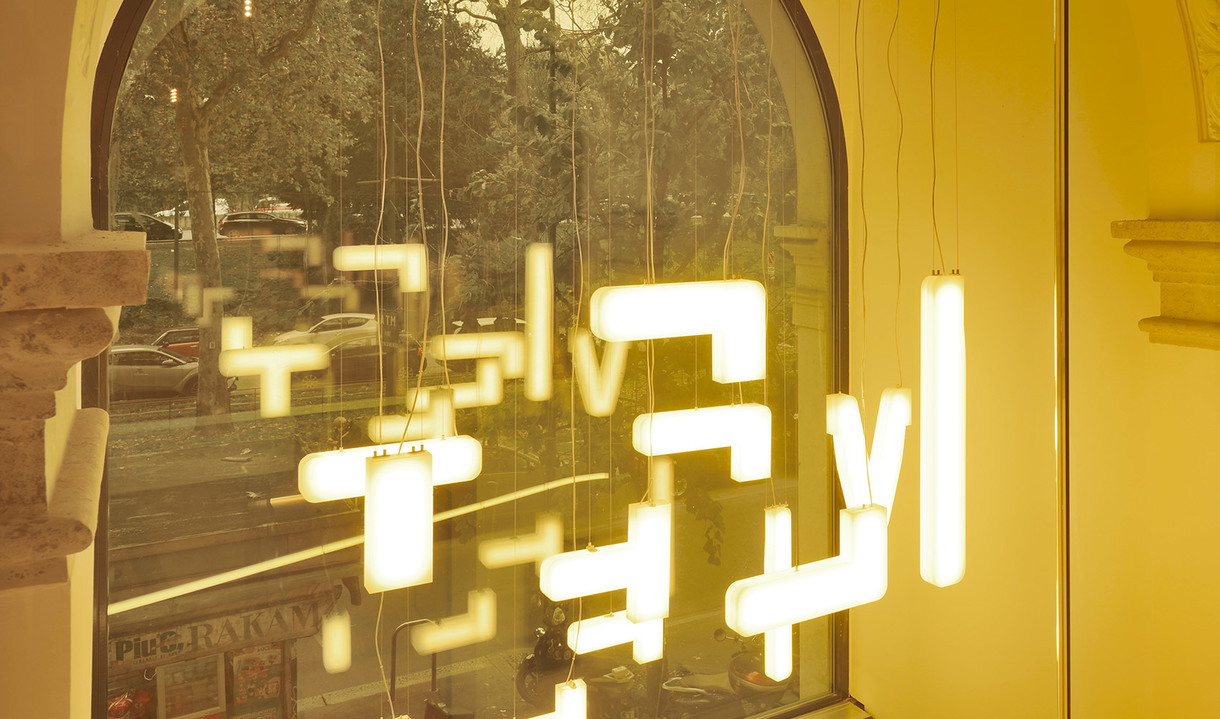 Multiple Artemide Alphabet of Light pendant modules arranged in geometric letter shapes, glowing warm white, suspended in an arched window display