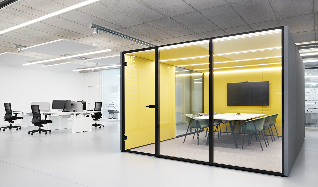 Ufficio moderno con postazioni aperte e una sala riunioni in vetro con pareti gialle, luci LED lineari a soffitto e tavolo conferenze.