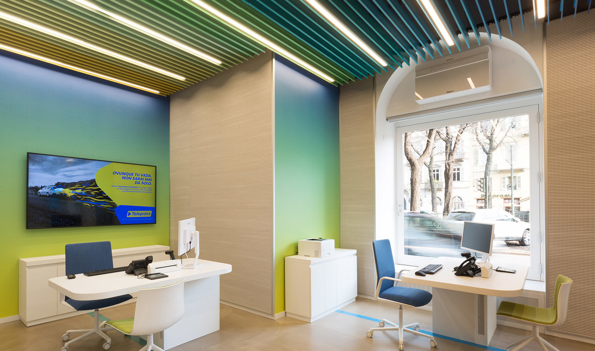 Interno di ufficio moderno con lampade lineari a soffitto Artemide, pareti con gradiente verde-giallo, scrivanie bianche e sedie blu.