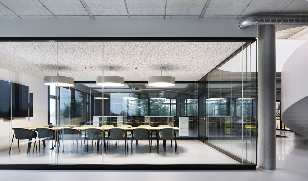 Sala conferenze moderna con pareti in vetro, lungo tavolo, sedie verdi e due lampade a sospensione cilindriche bianche.