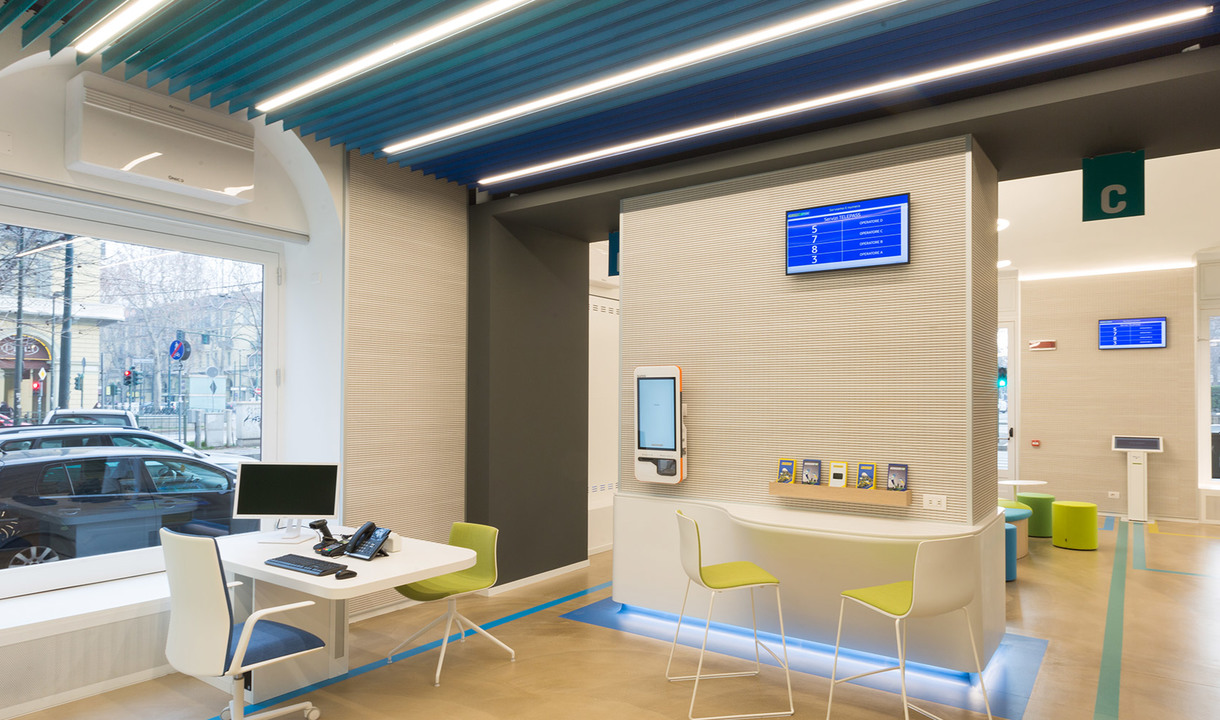 Interno moderno di banca con lampade lineari LED Artemide a soffitto che illuminano un'area servizi luminosa con postazioni e display digitali