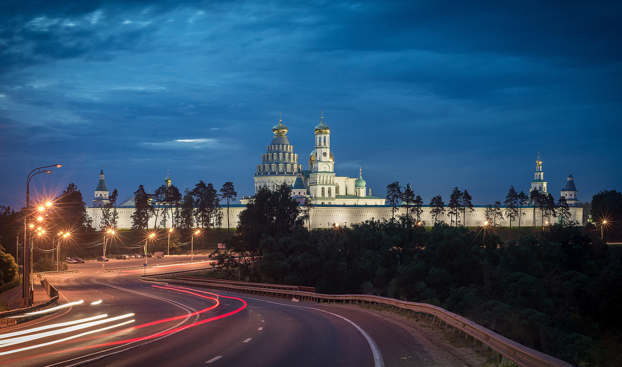 New Jerusalem Monastery illuminated at dusk, with light trails from passing cars on a curved road lined with street lamps
