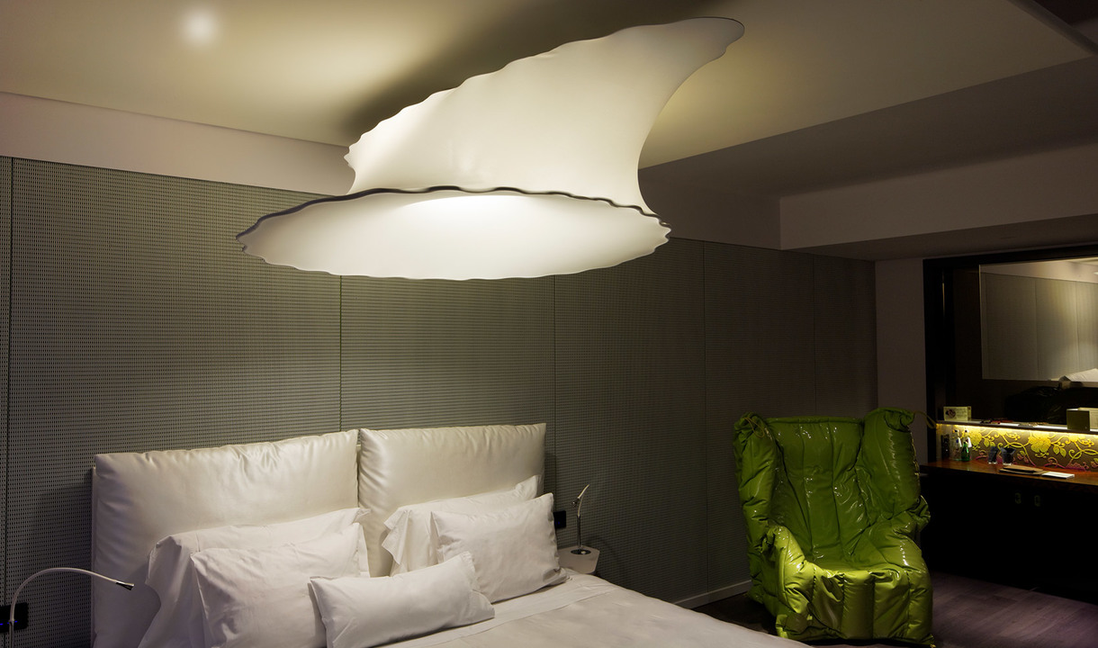 Grande lampada a sospensione Artemide a forma di nuvola che illumina una camera da letto moderna con biancheria bianca e poltrona verde.